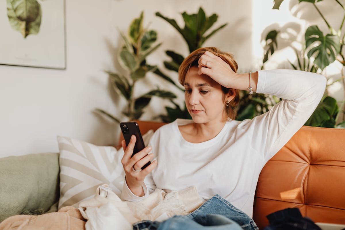 A woman in a white tee on a sunlit sofa focused on her phone, surrounded by indoor plants